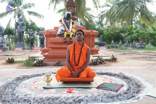 A seated man in saffron-orange robes sits cross-legged with eyes closed and hands folded in meditation on a low orange platform atop a rangoli-decorated circle of grey ash. He wears a long rudraksha mala and a red bindi on his forehead. Behind him stands a large orange-brick peetha with a black Shiva linga draped in a white and orange shawl and wrapped in yellow and purple garlands. Stone deity statues, palm trees and potted plants surround the courtyard. A brass kalasha sits to his left, and a small maroon cloth lies to his right.