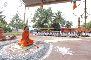 Outdoor satsang under a metal-roofed open pavilion with coconut palms behind. A man with long dark hair and a short beard sits cross-legged on a raised orange-cushioned seat on a mat, wearing an orange robe and a dark rudraksha mala, hands folded in his lap with eyes closed. In front of him is a brass kalasha-style vessel and a red flower on white rangoli patterns drawn on the ground. Dozens of seated people in white clothing face him, with mats laid out in the foreground. Colourful striped pillars and orange cloth decorations hang from the pavilion poles. Swamiji (probable): long hair, orange saffron robe and rudraksha mala, seated as teacher on the dais and leading the gathering.