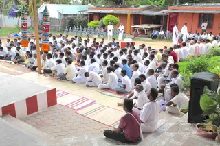 A large outdoor gathering of devotees seated on mats on a sandy courtyard, most wearing white dhotis and shirts, with some in coloured shirts. Many sit in rows facing a raised dais at the top left, while a few men stand near the back in white clothes. Decorated pillars with painted red, orange and blue panels frame the left side, and a black speaker sits at the right edge. Kolam-style white line designs are drawn on the ground and steps, and orange-roofed buildings with a flower garland hang in the background. Two standing figures in white near the centre appear to be attendants or priests; no one in the scene shows clear saffron or ornate cues for a person, so the figure is not confidently identifiable.