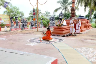 Outdoor temple courtyard with palm trees and a row of grey stone sculptures of deities and figures on low plinths. In the centre, a person in orange robes and a rudraksha mala sits cross-legged on a mat within a circular ring of ash, with a small brass vessel and a lamp before them, hands raised in a gesture of blessing. Two men in white dhoti and shawl stand on the right near a large dark stone Nandi bull with a flower garland, beside a tall orange and grey shrine pillar. A bamboo mat runs across the foreground over a patterned white kolam design on the ground. A yellow banner with printed text is on the left.