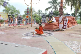 Outdoor temple courtyard with red-brown plinths and grey stone sculptures of deities and serpent hoods along the left. A figure in orange robes and a rudraksha mala sits cross-legged on a raised orange platform beside a small brass lamp inside a sand-coloured circular kolam. In the background on a larger plinth, a seated black stone Nandi-like bull figure wears a garland, and two men in white dhoti and shawl stand nearby, one holding a staff, the other with hands folded. Palm trees and a tall orange-and-white ritual pole frame the scene, and a banner with a portrait is at the left edge. A woven mat runs across the ground toward the platform.