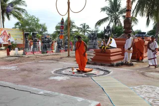 Outdoor temple courtyard with palm trees and a sandy ground. In the centre, a man in saffron-orange robes and a rudraksha mala stands on an orange mat within a stone-rimmed circle decorated with white rangoli lines, a small brass vessel in front of him. Behind him, a large black stone Shiva lingam sits on a red pedestal, adorned with white cloth and yellow-orange garlands, and several dark stone deity sculptures stand on red plinths. Two men in white dhoti and shawl walk nearby and another man in orange robes stands at right, with a bright banner at left showing printed figures. A woven mat runs across the ground toward the foreground.