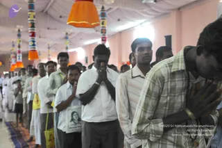 A photograph of a long line of men standing in a hall with white fabric ceiling draped overhead and tall striped decorative poles with hanging orange and yellow fabric panels. In the foreground a man in a white shirt and grey trousers presses his palms together in anjali mudra with eyes lowered, while a man in a plaid shirt bows his head with hands folded. A man in a white shirt holds a framed printed portrait of a woman and a yellow tote bag hangs from another man's arm. Other devotees in white shirts and dhotis stand behind them in an orderly queue along a purple and orange carpet. Several participants appear to be in a darshan or satsang line.