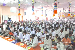 A large indoor hall filled with hundreds of seated devotees on orange and red mats, facing a stage at the front. Most wear white shirts and white dhotis or pants, with some in colourful shirts and saris. Tall cylindrical decorative pillars in patterned yellow, blue, red and white hang from a white ceiling canopy. On the left, a flat-screen monitor displays a seated figure in orange, beside a video camera on a tripod. Many attendees hold yellow notebooks and papers, and some wear lanyard badges.