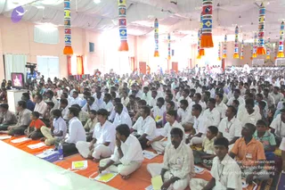 A large indoor satsang hall filled with hundreds of seated attendees on orange-and-white mats, most men in white shirts and dhotis or trousers, some in lighter or colored clothing, and a few children in front. Attendees sit facing a stage at left, where a small monitor shows a figure in red-orange clothing. Colorful cloth-wrapped pillars with tassels hang from a white ceiling, and white drapes and orange curtains frame the hall. A camera on a tripod records the event. The crowd is densely packed across the room.