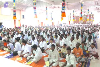 A large hall filled with a dense audience of men and boys seated on orange and white mats, most dressed in white shirts and white dhotis, with some wearing orange or colourful shirts. Many hold yellow and white booklets and wear lanyard badges. Tall decorative pillars wrapped in coloured woven patterns with orange tassels hang from a white draped ceiling. At the far end, a stage area is visible beyond the crowd, and a video camera on a tripod stands at the left edge.