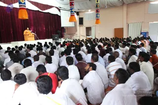 A large hall packed with seated devotees, mostly men in white shirts and dhotis, facing a raised stage. On the stage, a figure in saffron-orange robes sits behind a wooden podium with a microphone, appearing to give a talk. Maroon drapes form the backdrop, with tall orange and multicoloured tasselled banners hanging on either side and a projector screen above. Several women in saris sit at the back, and a camera operator is visible on the right. a person (orange robes at a discourse podium) delivering the satsang discourse to the assembly.