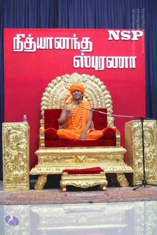 A figure in saffron-orange robes and an orange turban sits on an ornate gold throne with red velvet cushions, a gold footstool before it, wearing a long strand of gold-beaded necklace and a red tilak on the forehead. He raises one hand while speaking into a microphone on a stand at right. Behind him is a red backdrop with white Tamil lettering and a dark blue curtain. Gold-carved pedestals flank the throne, and a water bottle sits on the left pedestal. The stage has a patterned rug.