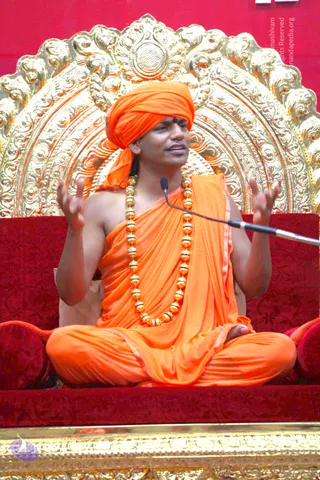 A seated man in saffron-orange clothing speaks into a microphone while gesturing with both raised hands. He wears an orange turban with a small red tilak mark on the forehead, a saffron shawl draped over one shoulder, and a long necklace of large gold beads. He sits cross-legged on a red upholstered seat in front of an ornate gold-embossed backrest with floral and scrollwork patterns. The page associates the event with a Tamil morning satsang talk; a swami is present in the photograph, with appearance cues of saffron clothing, a turban and gold beads, so the identity is marked as probable.