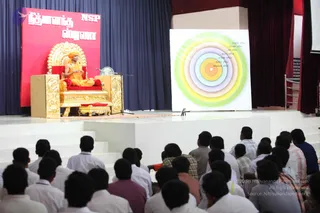A seated figure in saffron-orange robes and an orange turban sits on an ornate golden throne with red cushions on a stage, speaking into a microphone. A red banner behind him reads Nithyananda NSP in Tamil and English. To his right stands a large white board with concentric coloured rings of green, yellow, pink, lavender and blue around a central orange circle, with small Tamil labels along the edge. Rows of seated audience members, many in white shirts, face the stage from the front, with more people on the right. A dark blue curtain and maroon drapes frame the hall.