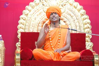 A seated man in saffron-orange robes and matching orange turban sits on a red cushioned throne before an ornate gold arched backdrop with a red wall behind him. He wears a long gold bead necklace, has a red tilak mark on his forehead, and raises his right hand in a teaching gesture while speaking into a microphone on a stand. A small plastic water bottle and a gold-trimmed side table appear at the left edge. The mood is that of a seated discourse.