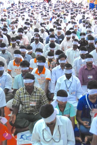 A large crowd of seated men and some women, many wearing white shirts and blindfolded with white cloth strips, fills the frame in a meditative gathering. Many wear identification badges and rudraksha-style beaded necklaces. A few wear orange-trimmed white polo shirts printed with N.D.S. lettering. Some sit cross-legged on an orange cloth floor while others sit with hands resting in their laps.