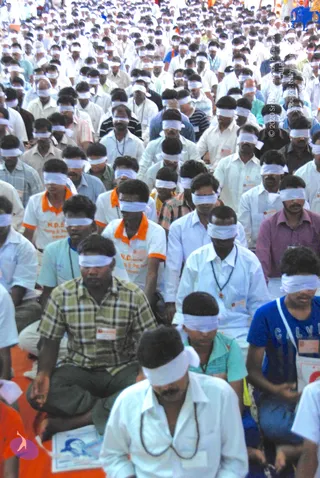 A very large crowd of seated devotees, mostly men in white shirts and light clothing, all wearing white blindfolds over their eyes during a meditation session. Many wear rudraksha-style beaded necklaces. Some have orange-collared white polo shirts printed with N.D.S. lettering. Several participants wear identification badges. Some sit cross-legged on an orange-toned floor covering, and a small printed portrait sheet lies in the lower left foreground.