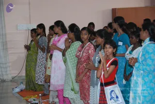 A photograph of a standing row of women and girls in an indoor hall with pale pink walls, a wall socket and a wooden cabinet at upper right. Most have palms pressed together in namaste at chest height. A girl in the foreground wears a red top and black skirt and carries a white tote bag printed with a portrait of a smiling woman. Others wear saris and salwar kameez in green, pink, blue, white and yellow tones, and some have name badges clipped to their clothes. An orange mat lies on the floor at lower left. No a person candidate is visible.