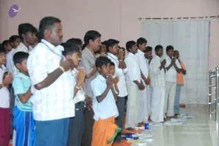 A photograph of a line of men, boys and youths standing with palms pressed together in namaste in an indoor hall with pale pink walls and a sheer white curtain on a rod. Men in white shirts and white dhotis stand near the front, a boy in an orange-and-green dhoti kneels in front, and a boy in a striped teal shirt and blue lungi stands at the left. Several participants wear white shirts; one older man wears a checked blue-and-white shirt. A steel rack stands at the right edge, with a water bottle and folded papers on the glossy floor. No visible a person figure is present; the scene shows devotees at a satsang-style gathering.