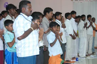 A photograph of a group of men and boys standing and kneeling in a hall with pink walls and a white curtain, all with hands joined in namaskar. In the foreground a man in a light blue checked shirt, blue trousers and a gold bracelet stands with palms pressed together, with a dark rudraksha-style mala visible at his neck. Beside him a boy in a white shirt and orange dhoti kneels with folded hands, and other boys in white shirts and dhotis stand behind them.