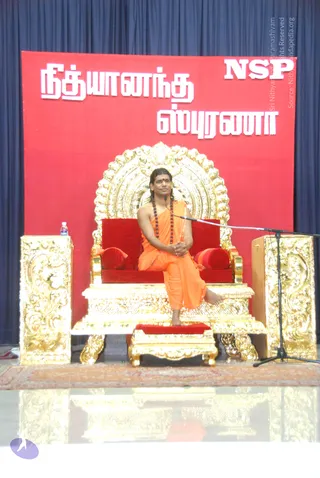 A photograph of a long-haired man in saffron-orange robes and a rudraksha mala seated on an ornate gold-and-red throne on a stage. He sits cross-legged with one foot on a gold footstool, hands clasped in his lap, and a microphone on a stand is angled toward him. Behind him a red banner with white Tamil lettering and the letters NSP hangs in front of navy curtains, and a water bottle rests on a gold side pedestal.
