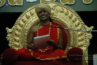 A seated man in red and gold ceremonial dress sits cross-legged on an ornate gold throne with curved, carved backrest and red cushions. He wears a light beige turban with a red bindi, a red shawl with gold border, and multiple gold-beaded necklaces. He holds an open white notebook or tablet in his hands and smiles while looking up to the side, with a microphone on a stand near him. Behind the throne, a banner shows large gold letters on a dark green background.