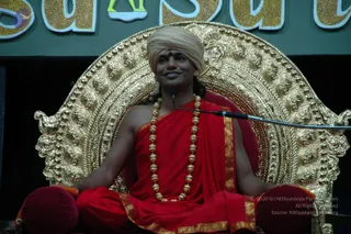 A seated man in a red saffron-toned robe with gold-bordered edges sits on an ornate gold throne with a red cushion, smiling. He wears a cream-coloured turban with a red bindi on the forehead and a long gold-beaded necklace, and his hands rest on red cushions at his sides. A microphone on a stand is positioned beside him. Above the throne, large gold-glittered letters on a green backdrop are partly visible.