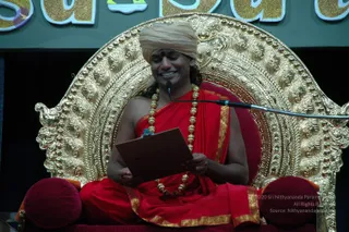 A smiling man sits on an ornate gold throne with a carved, scrolled back and a red cushion. He wears a peach-coloured turban, a red robe with gold border, and a long gold bead necklace. He holds a brown folder or booklet open in both hands and has a microphone on a stand beside him. Partial lettering in gold and blue is visible on the backdrop behind the throne.