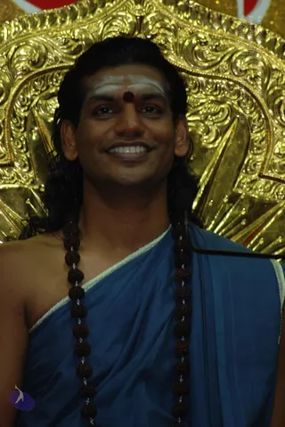 A smiling man with long dark hair worn back and a white vertical tilak with a red dot on his forehead sits before an ornate gold repoussé backdrop. He wears a blue draped cloth over his shoulder with a light border and a long necklace of dark rudraksha beads. A microphone stand is visible at the right edge. The presentation is consistent with Swamiji (probable) from the appearance cues: long swept-back hair, saffron-free but ceremonial ochre-style gold setting, and rudraksha mala, though the image is a close portrait.