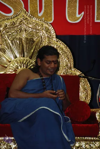 A man seated on an ornate gold throne with a red velvet cushion, wearing a blue draped garment with a silver-white edge and a dark rudraksha mala. He has long swept-back hair and a red bindi on his forehead, and holds a bead or rudraksha in his hands while smiling and looking down. A microphone stand is visible at right, and a red and gold banner with glittering letters is behind him. The person is an appearance-based probable candidate for Swamiji, with cues of long swept-back hair, rudraksha mala and ornate gold throne, but the identification is not certain.