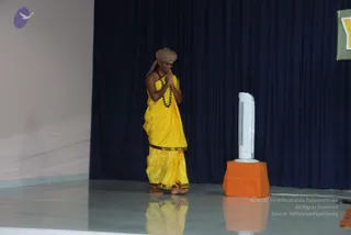 A man stands barefoot on a glossy grey stage with his palms pressed together in namaste, head slightly bowed. He wears a golden-yellow silk garment draped over his body with a dark gold border, a tan turban-like head wrap, and a dark beaded rudraksha-style mala around his neck. A navy blue curtain forms the backdrop, and a white tower fan stands on an orange-draped pedestal to the right. A green framed sign is partly visible at the top right edge. The stage floor reflects the figure.