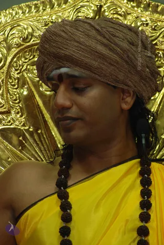 Close photo of a man seated before an ornate gold repoussé throne backdrop. He wears a brown crinkled turban, a red vertical mark and white ash on his forehead, and a yellow silk garment with a dark border. Long rudraksha mala beads hang on both sides of his chest, and a black microphone is clipped near his shoulder. His eyes are lowered and his expression is calm. Swamiji (probable): saffron-yellow clothing, turban and rudraksha malas are visible, and he sits as a seated speaker.
