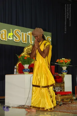 A photograph of a man on a stage standing barefoot with palms pressed together in namaste, eyes lowered. He wears a tan turban, a long yellow silk garment with gold-brown borders, and a dark rudraksha mala around his neck. Behind him is a golden seat with red cushions, a golden deity murti or ornament, and two vases of orange gerbera flowers with greenery on white pedestals. A large green banner with gold lettering stands behind. The man is one visible person.