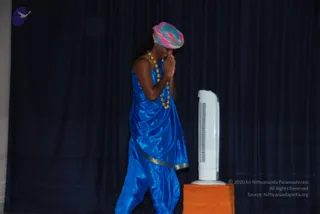 A performer stands on a stage before a dark blue draped curtain, hands pressed together in namaste at the chest with head bowed. He wears a pink, teal and green patterned turban, a long blue satin robe with a gold-edged hem and blue draped trousers, and a multi-strand gold and beaded necklace. Beside him a white tower fan stands on an orange-draped pedestal.