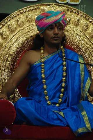 A man seated on an ornate gold throne with a red cushioned seat, wearing a multicoloured pink, teal and yellow turban with a red bindi on his forehead. He wears a royal blue silk garment with gold border, a gold bead mala around his neck, and gold bangles. A microphone stand is visible at right. Behind him is a partially visible gold lettered sign at top.