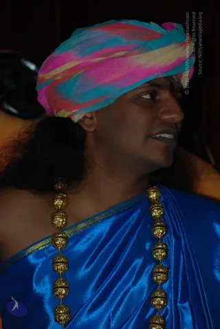 Close profile photograph of a man wearing a multicoloured pink, turquoise and yellow turban wrapped in a striped fabric, with a red tilak mark on his forehead. He has long dark hair falling behind his shoulder and wears a shiny royal blue silk garment edged with gold trim. Heavy gold beaded necklaces in large round ornamental beads hang over his chest. The background is dark with a blurred figure at the left edge. a person: the long swept hair, ornate gold necklaces and saffron-adjacent ceremonial styling are visible cues, appearing as an in-person subject.