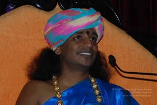A photograph of a man seated in front of an orange upholstered chair with a dark carved wooden frame and a microphone on a stand at the right edge. He wears a multicoloured pink, blue and cream turban with a red dot on his forehead, long dark hair falling past his shoulders, a blue satin shawl draped over one shoulder and a long gold bead necklace of ceremonial beads.