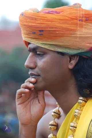 Close side profile photograph of a man wearing an orange and yellow tie-dyed turban with gold threads and blue dots, a red tilak mark on his forehead, and a yellow garment with gold embroidered border. His hand rests at his chin in a thoughtful pose, and he wears a heavy gold necklace of ornate beads. Blurred greenery and buildings fill the background. The visible cues are a turban, gold ceremonial beads and yellow-orange clothing, but the face is not clear enough for a confident candidate match, so the identity is uncertain.