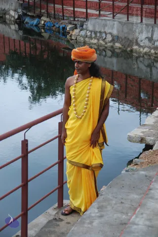 A man stands on a concrete ghat beside a still water tank, next to a red metal railing. He wears an orange-and-pink turban, a yellow draped garment with a dark gold-patterned border, and a long necklace of large gold-toned beads. His long dark hair falls past his shoulders and he wears open sandals. Red-painted steps and walls are reflected in the water behind him.