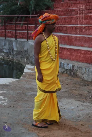 A man stands in profile on a dirt ground beside a shallow water channel and red brick steps with a railing and palm tree behind him. He wears a multicoloured orange, green and pink turban, a yellow draped garment with dark ornamental borders over one shoulder, and a long necklace of large gold beads. He is barefoot in black and orange sandals and holds his right arm relaxed at his side.