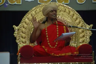 A man seated on an ornate gold throne with a red cushioned seat speaks at an event. He wears a cream turban with a red bindi, a red robe with gold border, and a long gold bead necklace. His right hand is raised in a gesture while he holds a white sheet of paper in his left hand. A microphone stand is at right, and a gold-lettered banner is visible at the top. Swamiji (probable): the appearance-based cues are the cream turban, red-and-gold robe and ornate gold beads, and he is seated on the throne as the speaker.