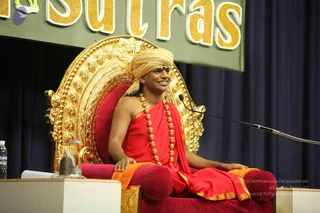A man seated on an ornate gold throne with a carved backrest and red cushions, smiling and turned to the side. He wears a saffron-yellow turban with a red bindi, a red robe with gold-trimmed edges, and a long strand of rudraksha beads with gold accents. A microphone on a stand is at his right, and a small silver shivalingam sits on a wooden table in front, beside a water bottle. Behind him hangs a dark blue curtain beneath a green banner with gold lettering reading 'Sutras'.