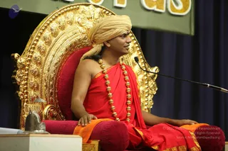 A seated man on an ornate gold throne with a red cushioned back and red-and-orange cushions. He wears a golden-beige turban, a red-and-gold draped robe with an orange shawl across the lap, and a long necklace of golden beads. His hand rests on the armrest, and a microphone on a stand is at right. Behind him are a dark blue curtain and a green banner with gold lettering. At lower left sits a small silver shivalinga on a white pedestal.
