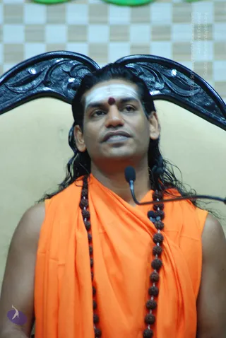 A man with long dark hair, a white forehead band and a red bindi sits in a carved black wooden chair in front of a checkered backdrop and speaks into a clip-on microphone. He wears an orange sleeveless wrap over a shoulder, and multiple rudraksha mala beads hang across his chest. The visible cues of long swept hair, orange clothing and rudraksha malas fit the a person candidate, and the page lists him at this event, so the identification is probable rather than certain.