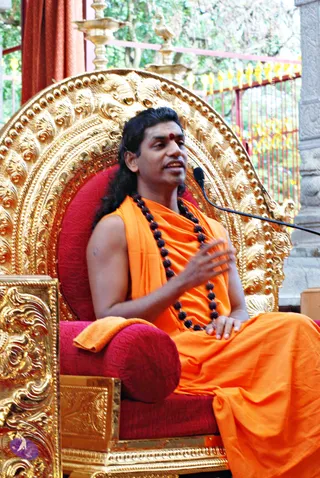 A man with long, swept-back dark hair and a red bindi sits on an ornate gold throne with a red cushion, speaking into a microphone on a stand. He wears an orange saffron wrap over a sleeveless orange garment and a long rudraksha mala. One hand rests on his lap while the other gestures in front of his chest. Behind him are red metal railings with gold finials, a gold kalasha-topped structure and green foliage. Swamiji (probable) is seated, with long swept-back hair, saffron clothing and a rudraksha mala, giving a morning message.