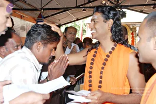 A photograph at an indoor darshan setting under a corrugated metal roof with ceiling fans and lights. A long-haired man in a sleeveless saffron-orange robe and a multi-strand rudraksha mala smiles while placing his hand on the head of a man in a white checked shirt who stands with palms pressed together in anjali. The robed man holds folded papers in his other hand. Behind them, several devotees in white and dark clothes watch, one with hands joined. A framed newspaper clipping and a colourful cloth hang on the left wall, and a gilded arch appears at the right.