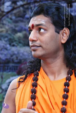 Close-up photograph of a man with long, dark, swept-back hair wearing an orange saffron shawl draped over his shoulders and a long rudraksha mala with large dark beads. He has a red bindi and white horizontal tilak marks on his forehead, and he looks off to the side with a thoughtful expression. The blurred background shows purple-tinged foliage.