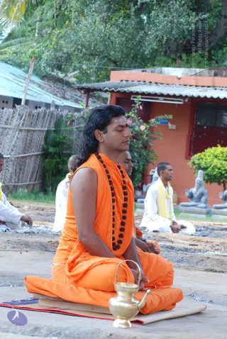 A man with long dark hair sits cross-legged with eyes closed in meditation on a low wooden seat over a red and green mat, outdoors on bare ground. He wears an orange sleeveless garment with a draped orange cloth and a long rudraksha mala with dark beads, and has a red tilak on his forehead. A brass vessel with a spout and handle sits in front of him. Behind him, other seated people in white clothes with yellow shawls meditate, and a row of grey stone deity murtis stands near an orange building with a corrugated roof.