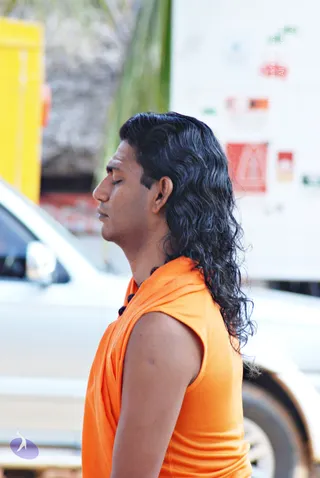 Side profile photo of a man with eyes closed in a standing meditative pose, long dark wavy hair falling past his shoulders, wearing a sleeveless orange saffron garment draped over one shoulder and a dark rudraksha-style bead mala at his neck. A white vehicle and a yellow structure sit blurred in the background, with a white wall displaying red and orange signage.