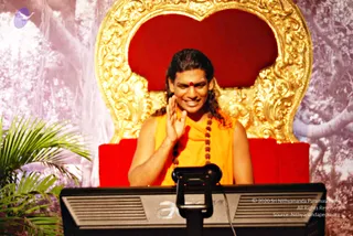 A man seated on a red and gold ornate throne before a forest-style backdrop of tree trunks and foliage, with a palm plant at left. He wears an orange-yellow sleeveless garment and a long rudraksha mala, has a red tilak on his forehead and long swept-back hair, and smiles while raising one hand in greeting. A black monitor stand with a lectern-mounted device sits in the foreground.