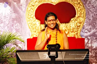 A man in a saffron-orange sleeveless garment sits on a red and gold ornate throne in front of a monitor, raising his right hand in a blessing gesture while smiling with eyes closed. He wears a red tilak on his forehead and a long mala of rudraksha beads, and his long hair is swept back. Green palm fronds stand at left against a backdrop of stylized forest trees.