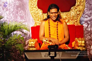 A smiling man with long dark swept-back hair and a red bindi sits on an ornate gold-and-red throne with carved golden lion heads at its base. He wears an orange saffron shawl over a bare upper body and a long rudraksha mala with a gold-toned trident-style ornament in his hands. A black audio monitor or stand is in the foreground, a palm plant stands to the left, and a forest-themed backdrop with pale foliage fills the background.