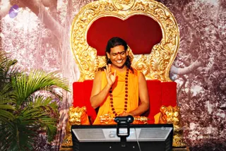 A man with long, swept-back dark hair sits on an ornate gold throne with a red cushioned seat and carved lion-head armrests, in front of a backdrop of a banyan-like tree in muted pink tones with a palm frond at left. He wears an orange sleeveless robe and a long rudraksha mala, with a red tilak on his forehead, and smiles while raising his right hand in greeting. A small golden trident-like staff rests on the throne arm in front of a black monitor or lectern. Swamiji (probable): the speaker's cues are long hair, saffron robes, a rudraksha mala and a throne seat.