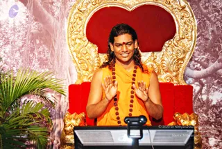 A man with long, swept-back dark hair sits on an ornate gold-and-red throne with carved lion-head armrests, in front of a stylised banyan-tree backdrop and a palm plant at left. He wears a saffron-orange sleeveless garment and a long rudraksha mala, with a red bindi on his forehead, and he gestures with both palms open toward a monitor and camera in the foreground. Swamiji (probable): long swept-back hair, saffron clothing and rudraksha mala, seated on a throne.